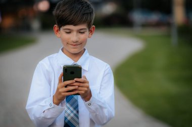 Beyaz gömlekli ve mavi ekose kravatlı genç bir çocuk şehir parkında yürürken akıllı telefonuna odaklanıyor. Arkaplan asfalt bir yol ve yeşillik gösteriyor