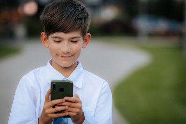Beyaz gömlekli ve mavi ekose kravatlı genç bir çocuk şehir parkında yürürken akıllı telefonuna odaklanıyor. Arkaplan asfalt bir yol ve yeşillik gösteriyor