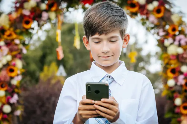 stock image A young boy dressed in a white shirt and blue plaid tie is smiling as he uses a smartphone. He stands outdoors, with a vibrant floral arch in the background, creating a festive and cheerful atmosphere