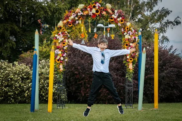 stock image A joyful young boy dressed in a white shirt and blue plaid tie leaps into the air with arms outstretched, smiling brightly. He is standing in front of a colorful floral arch with large pencil