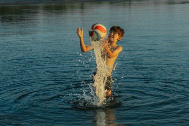 Genç bir çocuk sığ ve sakin denizde bir plaj topuyla oynuyor. Temiz gökyüzü ve su, sudaki neşeli anı vurgulayan sakin bir zemin sağlar. Uzayı ufkun üzerinde kopyala.