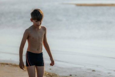 Genç bir çocuk sessiz bir plaj boyunca yalınayak yürüyor, aşağıya kuma bakıyor. Sakin su mesafeye kadar uzanıyor ve huzurlu bir manzara yaratıyor..