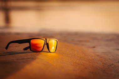 Kumda dinlenen bir güneş gözlüğü parlak turuncu bir günbatımını yansıtır. Aynalı lensler plaj sahnesinin sıcaklığını ve dingin atmosferini arttırır..