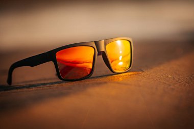 Kumda dinlenen bir güneş gözlüğü parlak turuncu bir günbatımını yansıtır. Aynalı lensler plaj sahnesinin sıcaklığını ve dingin atmosferini arttırır..