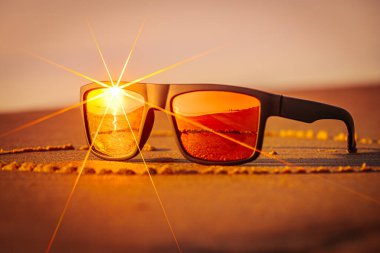Kumda dinlenen bir güneş gözlüğü parlak turuncu bir günbatımını yansıtır. Aynalı lensler plaj sahnesinin sıcaklığını ve dingin atmosferini arttırır..