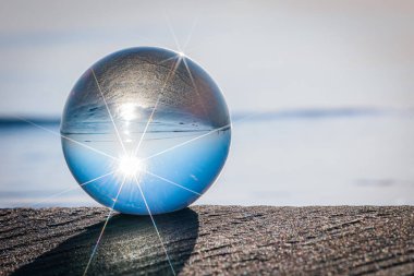 Açık bir cam küre kumsalda duruyor, mavi gökyüzünü ve plajı yansıtıyor, yıldız patlaması efektleriyle. Huzurlu arka plan kürelerin kristal berraklığını vurguluyor..