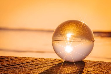 Cam bir küre kumlu bir sahilde, parlak bir yıldız patlaması etkisiyle parlayan günbatımını yansıtıyor. Gökyüzünün ve suyun altın rengi ılık ve huzurlu bir atmosfer yaratır..