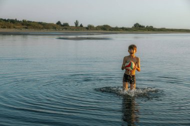 Genç bir çocuk sığ ve sakin denizde bir plaj topuyla oynuyor. Temiz gökyüzü ve su, sudaki neşeli anı vurgulayan sakin bir zemin sağlar. Uzayı ufkun üzerinde kopyala.