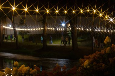 Bir grup insan gece bir parkta sıcak, parlak ip ışıkları altında toplanıyor. Şenlik ışıkları hoş bir ortam yaratıyor. 07.09.2024, Koceni, Letonya