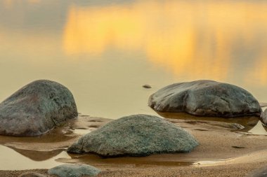 Kuma dağılmış kayalarla sakin bir sahil sahnesi ve batan güneşin altın renklerini yansıtan durgun bir su havuzu. Huzurlu deniz ufka doğru uzanıyor, barışçıl bir deniz yaratıyor.