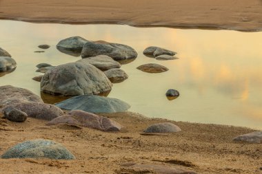 Kuma dağılmış kayalarla sakin bir sahil sahnesi ve batan güneşin altın renklerini yansıtan durgun bir su havuzu. Huzurlu deniz ufka doğru uzanıyor, barışçıl bir deniz yaratıyor.