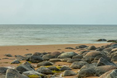 Kumun üzerine dağılmış kayaların olduğu sakin bir sahil sahnesi batan güneşin ılık renklerini yansıtan durgun bir su havuzuna yansıyor. Sakin deniz ve yumuşak kum huzurlu bir atmosfer yaratır..