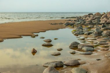Kumun üzerine dağılmış kayaların olduğu sakin bir sahil sahnesi batan güneşin ılık renklerini yansıtan durgun bir su havuzuna yansıyor. Sakin deniz ve yumuşak kum huzurlu bir atmosfer yaratır..