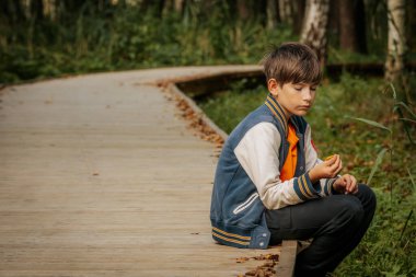 Genç bir çocuk bir ormanda ahşap bir patikada oturur, elinde bir nesneyle dalgın dalgın aşağıya bakar. Düşen yapraklar patika boyunca uzanır, sonbahar dokunuşu ekler..