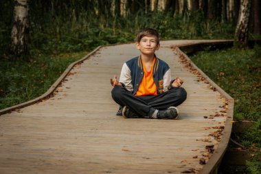 Genç bir çocuk tahta bir yolda bacak bacak üstüne atarak oturur, gözleri kapalı meditasyon yapar, sakin bir ormanda sakin ve düşünceli bir anı kucaklar..