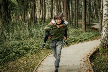 Neşeli genç bir çocuk, sakin bir orman ortamında ağabeyinin sırtında gezintiden zevk alırken gülümsüyor. Arka plandaki uzun ağaçlar doğal ve huzurlu bir atmosfer ekler..