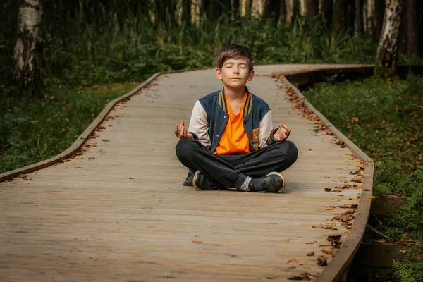 Genç bir çocuk tahta bir yolda bacak bacak üstüne atarak oturur, gözleri kapalı meditasyon yapar, sakin bir ormanda sakin ve düşünceli bir anı kucaklar..