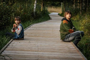 Genç bir çocuk ve yaşlı bir genç yeşil alanlarla çevrili ahşap bir patikada oturuyorlar. Genç çocuk bir çim sapı tutarken, genç düşünceli görünüyor, mesafeye bakıyor..