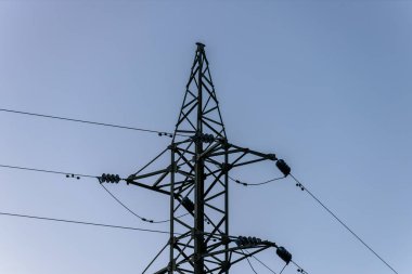 Açık mavi gökyüzüne karşı dimdik duran yüksek voltajlı bir elektrik hattı kulesi. Çelik yapı ve elektrik kabloları, arka planda doğal manzara ile çarpıcı bir endüstriyel kontrast oluşturuyor..