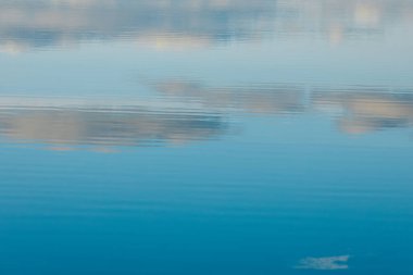 Augstroze Gölü 'nün sakin manzarası. Önünde durgun mavi sular ve uzakta ağaçlarla kaplı bir sahil. Ağaçlar açık bir gökyüzünün altında sonbaharın başlarında renkler sergilerler..