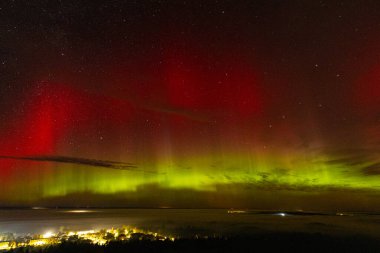 Çarpıcı kırmızı ve yeşil renkleri olan canlı bir Aurora Borealis bir kasabanın üstündeki yıldızlı gece gökyüzünü aydınlatıyor. Canlı göksel görüntü, aşağıdaki aydınlık kasaba Zilaiskalns ile güzel bir tezat oluşturuyor..