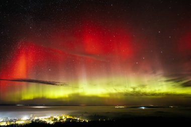 Çarpıcı kırmızı ve yeşil renkleri olan canlı bir Aurora Borealis bir kasabanın üstündeki yıldızlı gece gökyüzünü aydınlatıyor. Canlı göksel görüntü, aşağıdaki aydınlık kasaba Zilaiskalns ile güzel bir tezat oluşturuyor..