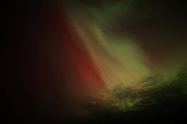 Aurora Borealis 'in canlı bir görüntüsü, koyu bulutlar arasında harmanlanmış kırmızı, turuncu ve yeşil renkleri ile gece gökyüzünü aydınlatıyor. Dramatik zıtlık gerçeküstü ve büyüleyici bir sahne yaratır..