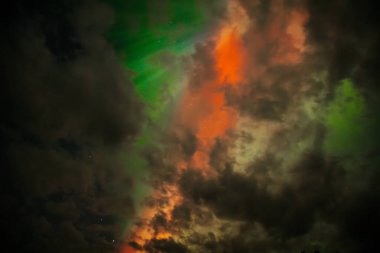 Aurora Borealis 'in nefes kesici görüntüsü gece gökyüzünü canlı kırmızı ve yeşil renklerle aydınlatıyor. Kara bulutlar kısmen karanlık Işıldayan ışıklar