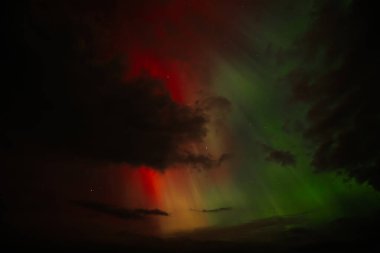 Aurora Borealis 'in canlı bir görüntüsü, koyu bulutlar arasında harmanlanmış kırmızı, turuncu ve yeşil renkleri ile gece gökyüzünü aydınlatıyor. Dramatik zıtlık gerçeküstü ve büyüleyici bir sahne yaratır..