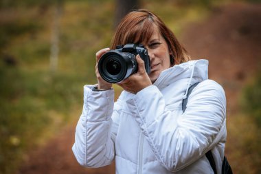 Beyaz ceketli bir kadın elinde DSLR kamerasıyla orman ortamında bulanık arkaplanlı bir fotoğraf çekiyor..