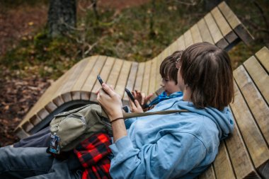 İki kardeş ormandaki ahşap bir bankta oturuyorlar, ikisi de akıllı telefonlarına odaklanmış. Açık havanın tadını çıkarırken rahat görünüyorlar..