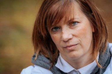 Uzun saçlı ve omzunda dövmesi olan bir kadın güneşli bir orman yolunda dikilirken sıcak bir şekilde gülümsüyor. Beyaz kolsuz bir bluz ve arkasında ağaçlar ve yeşillik olan kırmızı bir kolye giyiyor.