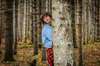 Siyah ceketli ve kışlık şapkalı bir çocuk karlı bir ormanda bir ağaç gövdesinin arkasından, doğal ortamda tuhaf ve neşeli bir hava yaratıyor..