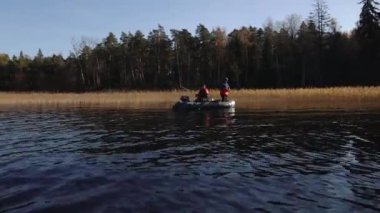 Mavi göldeki bir teknede balıkçılar, sakin bir günde olta atıp balık yakalıyorlar. Su sonbahar manzarasını yansıtır, dinginliği ve mevsimsel hissi arttırır. 10.19.2024, Letvija Limbai
