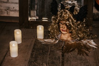 Kıvırcık saçlı, siyah boynuzlu, LED mumlarla çevrili bir manken kafası ve tahta bir yüzey üzerinde siyah bir fener içeren ürkütücü bir düzenleme. Loş, karamsar ışık rahatsız edici bir atmosfer yaratır..