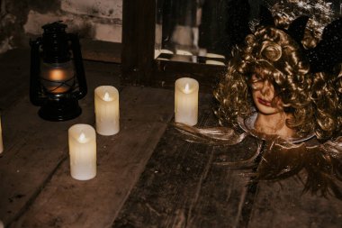 Eerie arrangement featuring a mannequin head with curly hair and black horns, surrounded by LED candles and a black lantern on a wooden surface. Dim, moody lighting creates a haunting atmosphere. clipart