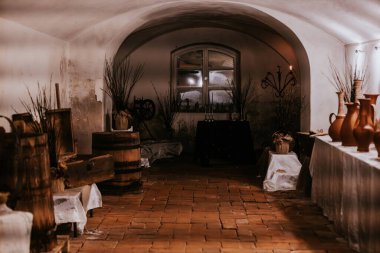 Antik dekorları, kil çömlekleri, ahşap fıçıları ve kurumuş dalları olan kemerli bir kiler odası. Yumuşak, karamsar ışıklandırma tarihi ve kırsal atmosferi güçlendirir..