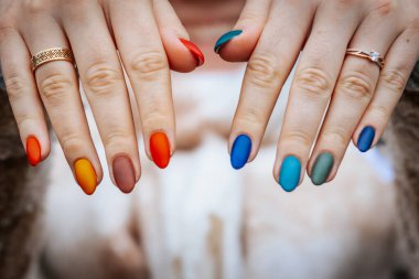 Bir kadının elleri kırmızı, turuncu, sarı, yeşil, mavi ve deniz mavisi tonlarında mat renkli manikür gösteriyor. Elleri iki yüzükle süslenmiş, biri elmas yüzük..