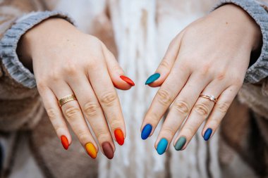 Bir kadının elleri kırmızı, turuncu, sarı, yeşil, mavi ve deniz mavisi tonlarında mat renkli manikür gösteriyor. Elleri iki yüzükle süslenmiş, biri elmas yüzük..