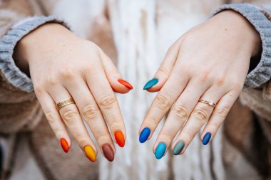 Bir kadının elleri kırmızı, turuncu, sarı, yeşil, mavi ve deniz mavisi tonlarında mat renkli manikür gösteriyor. Elleri iki yüzükle süslenmiş, biri elmas yüzük..