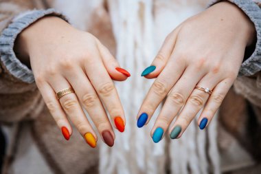 Bir kadının elleri kırmızı, turuncu, sarı, yeşil, mavi ve deniz mavisi tonlarında mat renkli manikür gösteriyor. Elleri iki yüzükle süslenmiş, biri elmas yüzük..