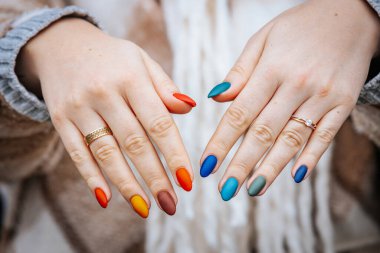 Bir kadının elleri kırmızı, turuncu, sarı, yeşil, mavi ve deniz mavisi tonlarında mat renkli manikür gösteriyor. Elleri iki yüzükle süslenmiş, biri elmas yüzük..