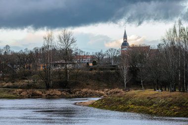 A peaceful village landscape by the riverside, featuring a church tower rising above bare trees. The cloudy sky adds a moody atmosphere, while the river curves gently. clipart