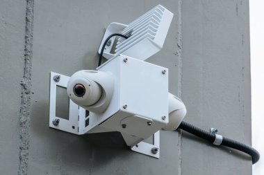 Close-up of a white, modern surveillance camera securely mounted on a wall. The camera features a sturdy bracket and connected wiring, designed for outdoor monitoring and enhanced security. clipart