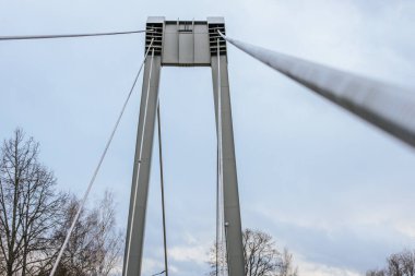 A low-angle view of a modern suspension bridge, showcasing the structural beams and cables. The bridge spans a natural landscape with tall evergreen trees and a cloudy sky in the background. clipart