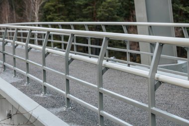 Modern asma köprünün alçak açılı görüntüsü, yapısal kirişleri ve kabloları gösteriyor. Köprü, arka planında bulutlu gökyüzü ve yüksek yeşil ağaçlarla doğal bir manzara boyunca uzanır..