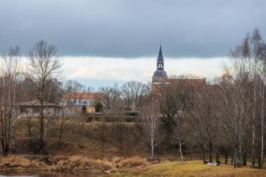 Nehir kenarındaki huzurlu köy manzarasında çıplak ağaçların üzerinde yükselen bir kilise kulesi var. Bulutlu gökyüzü karamsar bir atmosfer ekler, nehir yavaşça kıvrılır..