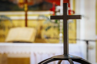 An iron cross stands prominently in the foreground with a blurred view of an open Bible and church altar in the background. The scene reflects a solemn atmosphere of faith, prayer, and worship inside  clipart