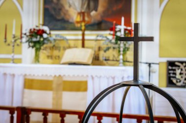 Arka planda açık bir İncil ve kilise sunağının bulanık görüntüsüyle ön planda belirgin bir demir haç duruyor. Sahne, inancın, duanın ve ibadetin kutsal atmosferini yansıtıyor. 
