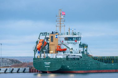 A large red cargo ship named Bugoe docked at a port with visible lifeboats and piles of woodchips under a cloudy sky. Ideal for maritime, trade, and industrial themes. 11.19.2024. Salacgriva,Latvia clipart